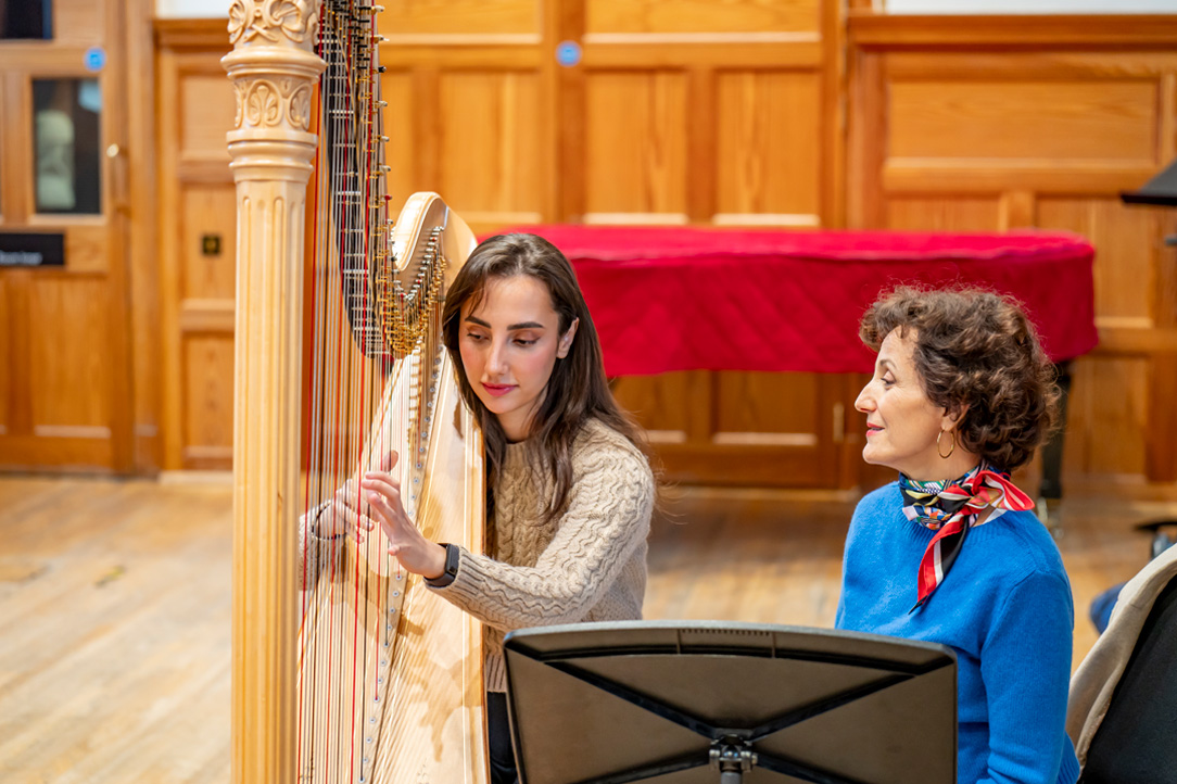 A female student playing the harp, with a women guiding her in a class, in a wood panelled room.